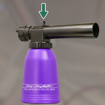 Kuvassa on violetin värinen spraypullomainen esine, nuoli osoittaa kohtaa josta painetaan alaspäin.